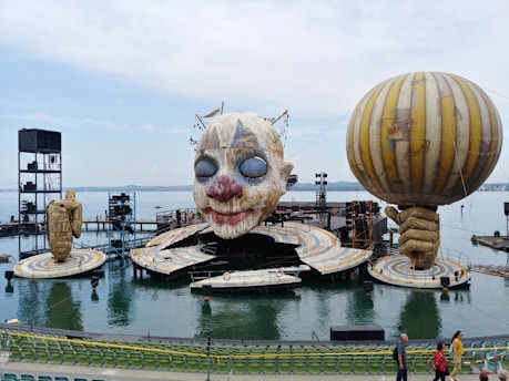 A large outdoor stage setup on a body of water features an oversized clown face. On either side of the stage, there are giant sculptural hands, one holding a balloon. The stage is surrounded by water and there is seating available in the foreground.