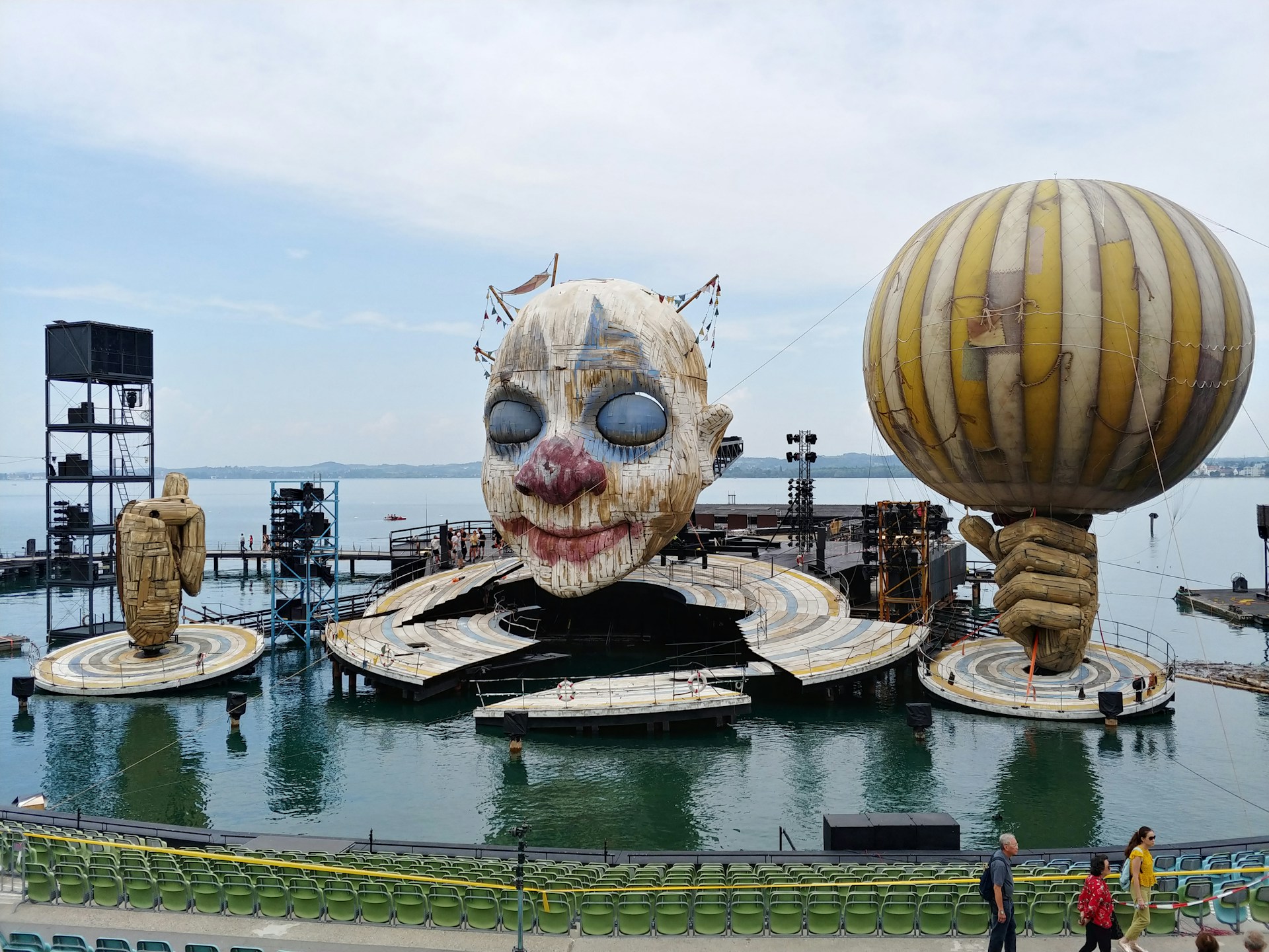 A large outdoor stage setup on a body of water features an oversized clown face. On either side of the stage, there are giant sculptural hands, one holding a balloon. The stage is surrounded by water and there is seating available in the foreground.