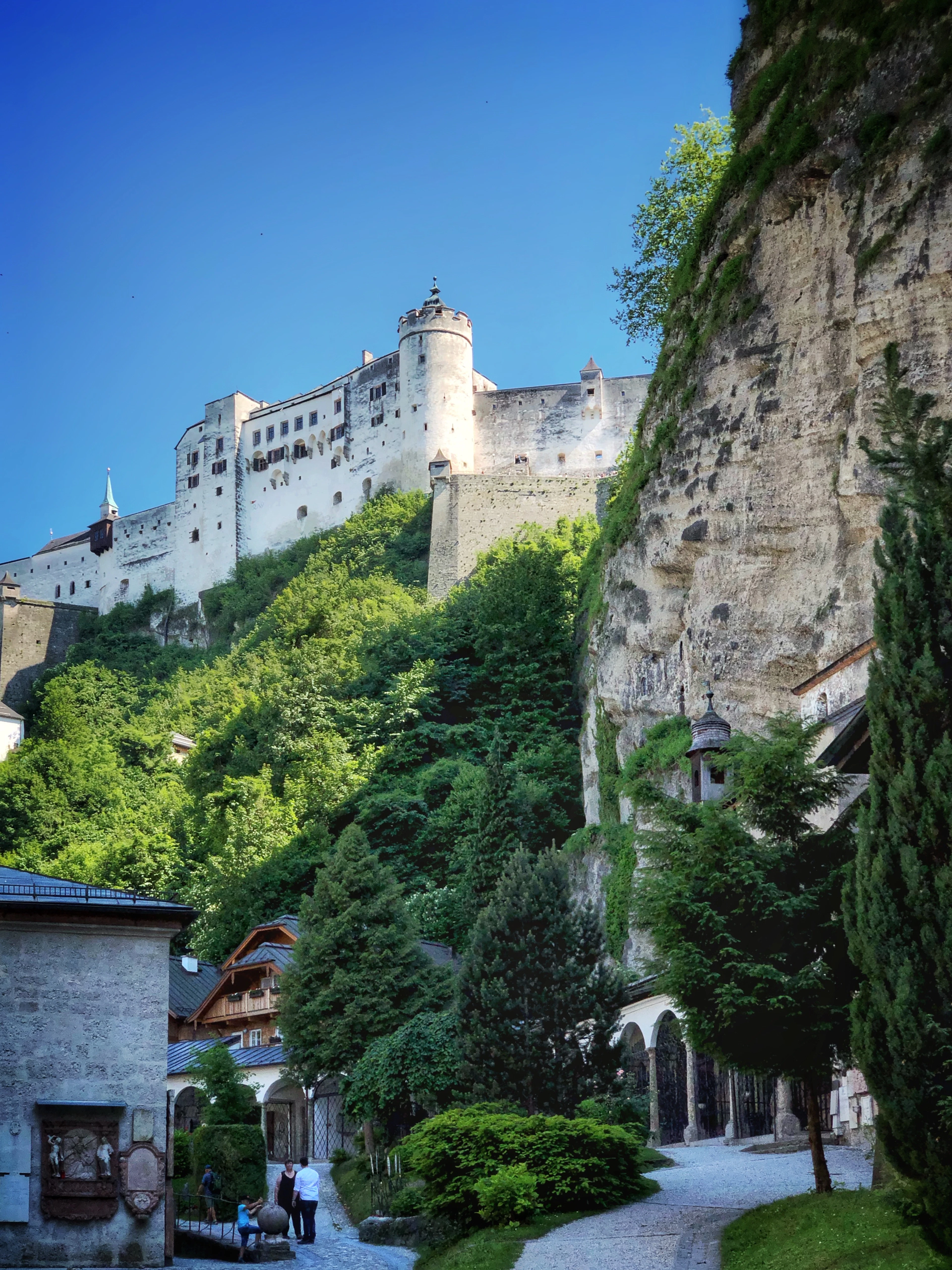 Hohensalzburg Fortress perched on a cliff above a quiet cloister walkway, seen on a clear blue-sky day.