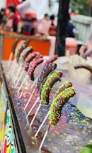 A row of chocolate-covered bananas on sticks is displayed on a table. Each banana is coated with colorful sprinkles. The background is blurred, featuring people and what seems to be a lively market or fair setting.