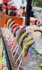 A row of chocolate-covered bananas on sticks is displayed on a table. Each banana is coated with colorful sprinkles. The background is blurred, featuring people and what seems to be a lively market or fair setting.