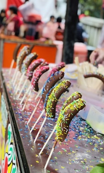 A row of chocolate-covered bananas on sticks is displayed on a table. Each banana is coated with colorful sprinkles. The background is blurred, featuring people and what seems to be a lively market or fair setting.