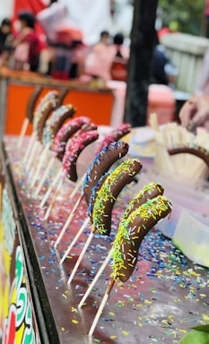 A row of chocolate-covered bananas on sticks is displayed on a table. Each banana is coated with colorful sprinkles. The background is blurred, featuring people and what seems to be a lively market or fair setting.