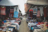 A market stall filled with various types of clothing, including shirts, pants, and underwear, displayed on tables and hanging on racks. The space is covered with a makeshift tarp, providing shade from above.