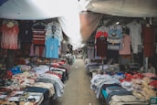 A market stall filled with various types of clothing, including shirts, pants, and underwear, displayed on tables and hanging on racks. The space is covered with a makeshift tarp, providing shade from above.
