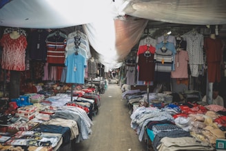 A market stall filled with various types of clothing, including shirts, pants, and underwear, displayed on tables and hanging on racks. The space is covered with a makeshift tarp, providing shade from above.