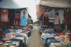 A market stall filled with various types of clothing, including shirts, pants, and underwear, displayed on tables and hanging on racks. The space is covered with a makeshift tarp, providing shade from above.