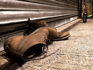 A pair of worn brown leather boots lies on a concrete sidewalk next to a closed metal storefront shutter. In the background, a bottle is visible standing upright a few feet away, suggesting an urban setting. The lighting casts a warm tone over the scene, emphasizing the texture of the boots and the roughness of the concrete.