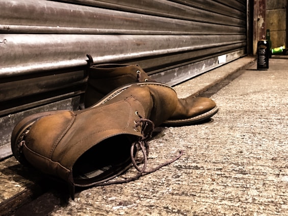 A pair of worn brown leather boots lies on a concrete sidewalk next to a closed metal storefront shutter. In the background, a bottle is visible standing upright a few feet away, suggesting an urban setting. The lighting casts a warm tone over the scene, emphasizing the texture of the boots and the roughness of the concrete.