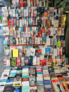 A large assortment of books is displayed on shelves in an outdoor setting, featuring a mix of Arabic and English titles. The books are arranged in rows and columns, showcasing a variety of genres, including fiction, non-fiction, history, and self-help.