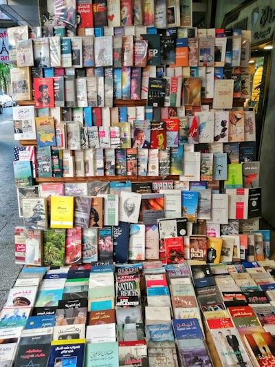 A large assortment of books is displayed on shelves in an outdoor setting, featuring a mix of Arabic and English titles. The books are arranged in rows and columns, showcasing a variety of genres, including fiction, non-fiction, history, and self-help.