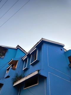 A vibrant blue building with white trim, showcasing geometric windows and balconies. The architectural design appears modern, with clean lines and minimalistic details. Visible wires contrast against the clear sky, and some greenery is present near the balconies.