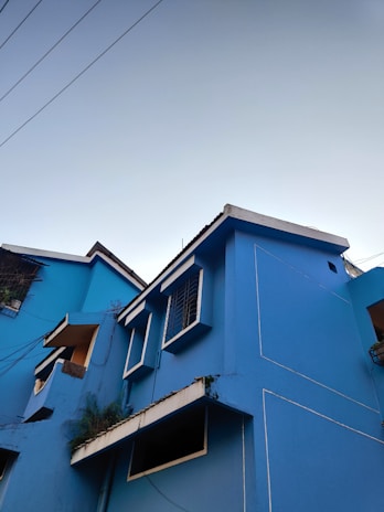 A vibrant blue building with white trim, showcasing geometric windows and balconies. The architectural design appears modern, with clean lines and minimalistic details. Visible wires contrast against the clear sky, and some greenery is present near the balconies.
