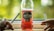 A bottle of Strongbow Red Berries apple cider is positioned in the center, surrounded by two hands holding lit cigarettes. The background appears to be a blurred natural setting, possibly with greenery, visible through a window.