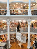 A spacious multi-level bookstore features floor-to-ceiling bookshelves filled with a diverse selection of books. The interior is marked by elegant architectural details, including tall white columns and a graceful spiral staircase connecting the levels. Several people are browsing the extensive collection, adding a lively yet tranquil atmosphere.