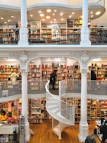 A spacious multi-level bookstore features floor-to-ceiling bookshelves filled with a diverse selection of books. The interior is marked by elegant architectural details, including tall white columns and a graceful spiral staircase connecting the levels. Several people are browsing the extensive collection, adding a lively yet tranquil atmosphere.