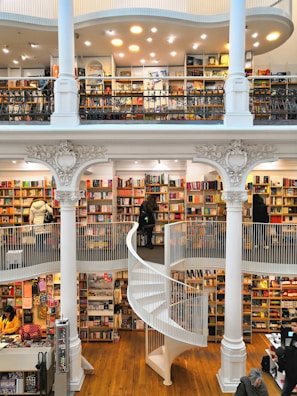 A spacious multi-level bookstore features floor-to-ceiling bookshelves filled with a diverse selection of books. The interior is marked by elegant architectural details, including tall white columns and a graceful spiral staircase connecting the levels. Several people are browsing the extensive collection, adding a lively yet tranquil atmosphere.