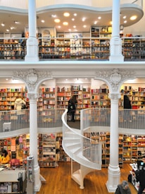 A spacious multi-level bookstore features floor-to-ceiling bookshelves filled with a diverse selection of books. The interior is marked by elegant architectural details, including tall white columns and a graceful spiral staircase connecting the levels. Several people are browsing the extensive collection, adding a lively yet tranquil atmosphere.