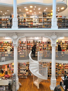 A spacious multi-level bookstore features floor-to-ceiling bookshelves filled with a diverse selection of books. The interior is marked by elegant architectural details, including tall white columns and a graceful spiral staircase connecting the levels. Several people are browsing the extensive collection, adding a lively yet tranquil atmosphere.