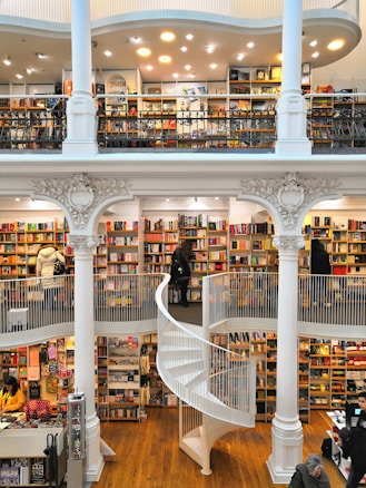 A spacious multi-level bookstore features floor-to-ceiling bookshelves filled with a diverse selection of books. The interior is marked by elegant architectural details, including tall white columns and a graceful spiral staircase connecting the levels. Several people are browsing the extensive collection, adding a lively yet tranquil atmosphere.