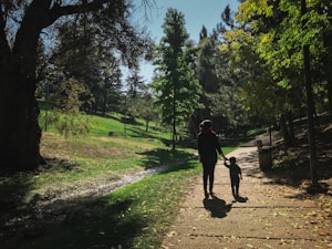 A peaceful park scene with a woman and a child walking hand in hand along a paved path. The surroundings are lush with green grass and tall trees, and sunlight filters through the leaves, creating dappled shadows on the ground. A small stream runs parallel to the path, adding to the tranquil atmosphere.