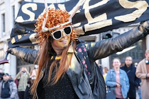 A person dressed in festive attire, featuring a bright orange wig and large sunglasses, spreads their arms wide in an expression of joy. The person is wearing a black leather jacket and stands in front of a banner with large gold letters. There are onlookers in the background.