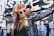 A person dressed in festive attire, featuring a bright orange wig and large sunglasses, spreads their arms wide in an expression of joy. The person is wearing a black leather jacket and stands in front of a banner with large gold letters. There are onlookers in the background.