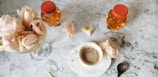 A marble table setting features a cup of coffee surrounded by delicate pink roses, two honey jars with red caps, and a decorative spoon.