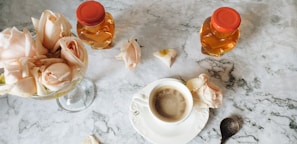 A marble table setting features a cup of coffee surrounded by delicate pink roses, two honey jars with red caps, and a decorative spoon.