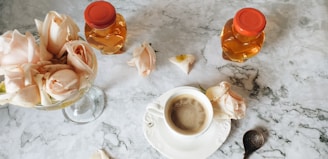 A marble table setting features a cup of coffee surrounded by delicate pink roses, two honey jars with red caps, and a decorative spoon.