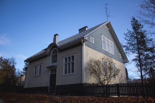 A two-story house with a steep gabled roof, painted in muted colors of blue and beige. It features multiple large windows and a chimney protruding from the roof. Bare trees surround the house, and a wooden fence encloses the property. The sky is clear and blue, indicating a calm day.