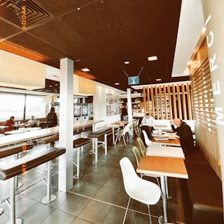 A modern indoor seating area of a fast-food restaurant features high stools and tables, with a few patrons sitting and eating. The room has a contemporary design with wooden accents and a tiled floor. Windows on one side allow ample natural light, while digital menu kiosks can be seen in the background.