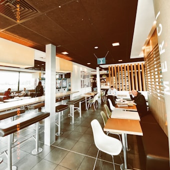 A modern indoor seating area of a fast-food restaurant features high stools and tables, with a few patrons sitting and eating. The room has a contemporary design with wooden accents and a tiled floor. Windows on one side allow ample natural light, while digital menu kiosks can be seen in the background.