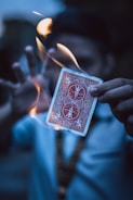 A person holds a playing card with a red back design as it is surrounded by swirling flames. The background is blurred, and the person's face is not in focus, emphasizing the card and the fire.