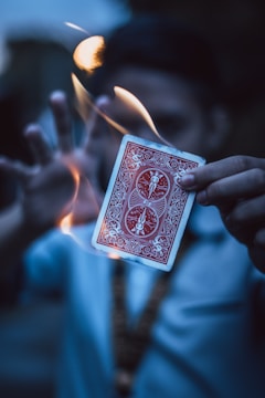 A person holds a playing card with a red back design as it is surrounded by swirling flames. The background is blurred, and the person's face is not in focus, emphasizing the card and the fire.