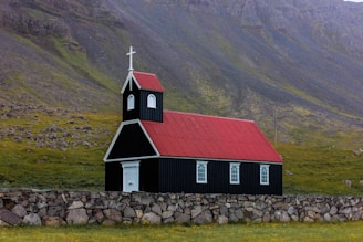 A small black church with a vibrant red roof is situated against a backdrop of rocky hills. The building features white-framed windows and a white door, topped with a steeple and cross. A stone wall surrounds the church, and green grass covers the ground.