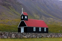 A small black church with a vibrant red roof is situated against a backdrop of rocky hills. The building features white-framed windows and a white door, topped with a steeple and cross. A stone wall surrounds the church, and green grass covers the ground.
