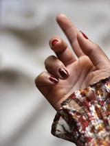 A hand with neatly manicured nails painted in a rich, glossy red color. The nails have intricate patterns, and the wrist is covered by a floral, patterned fabric in earthy tones.