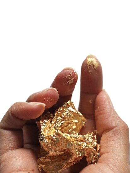 A hand is holding pieces of gold foil, with specks of the foil adhering to the fingertips. The background is a plain white, highlighting the contrast of the metallic shine.