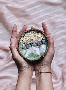 A pair of hands gently holds a coconut bowl filled with a green smoothie. The top of the smoothie is adorned with a neat arrangement of oats, fresh blueberries, and slices of coconut. The background has a soft, striped texture in shades of beige and light pink.