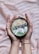 A pair of hands gently holds a coconut bowl filled with a green smoothie. The top of the smoothie is adorned with a neat arrangement of oats, fresh blueberries, and slices of coconut. The background has a soft, striped texture in shades of beige and light pink.