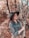 A person stands with closed eyes in a forest area, wearing a wide-brimmed hat and a patterned blouse. The background is filled with tall trees and autumn foliage, lending a rustic and serene atmosphere.