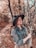 A person stands with closed eyes in a forest area, wearing a wide-brimmed hat and a patterned blouse. The background is filled with tall trees and autumn foliage, lending a rustic and serene atmosphere.