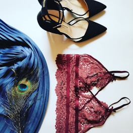 A neatly arranged flat lay featuring a pair of black high-heeled shoes with ankle straps, a burgundy lace bralette with intricate detailing, a pleated blue fabric with a peacock feather on top.