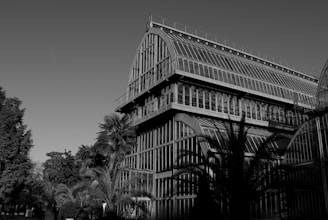 A large glass greenhouse structure with arched roofs, surrounded by various palm trees and foliage. The architecture features multiple levels with extensive glass coverage, allowing natural light to enter. The building is situated in a landscaped environment with additional greenery in the background.