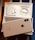 An opened box containing a smartphone and accessories. The smartphone, with a reflective back featuring a prominent logo, is placed on top of the box. Inside the box are neatly arranged earphones and a charging cable.