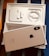 An opened box containing a smartphone and accessories. The smartphone, with a reflective back featuring a prominent logo, is placed on top of the box. Inside the box are neatly arranged earphones and a charging cable.