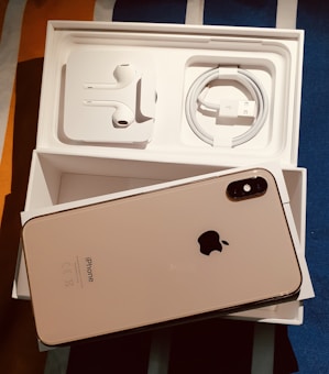 An opened box containing a smartphone and accessories. The smartphone, with a reflective back featuring a prominent logo, is placed on top of the box. Inside the box are neatly arranged earphones and a charging cable.