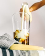 A hand is pouring milk from a glass bottle into a glass filled with coffee, creating swirls of cream in the dark liquid. A folded gray checkered towel is placed on a surface next to the glass.