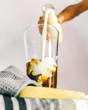 A hand is pouring milk from a glass bottle into a glass filled with coffee, creating swirls of cream in the dark liquid. A folded gray checkered towel is placed on a surface next to the glass.